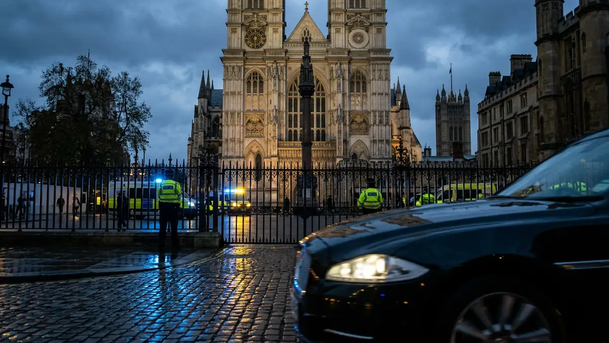 Westminster Abbey exterior at dusk with police light reflections on wet cobblestones.