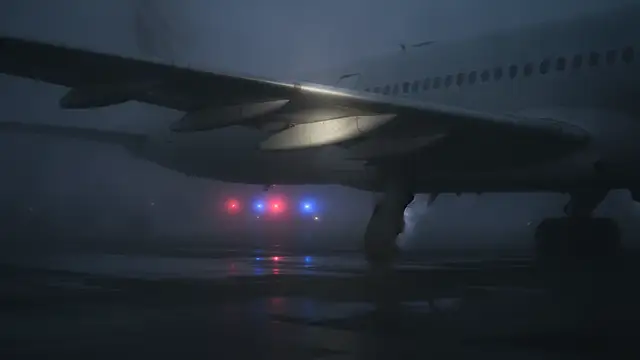 Aircraft wing shrouded in mist on tarmac, emergency lights reflecting.