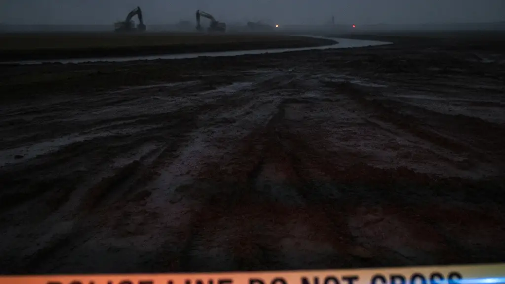 Police tape in foreground, blurred industrial machinery and muddy landscape near a river.