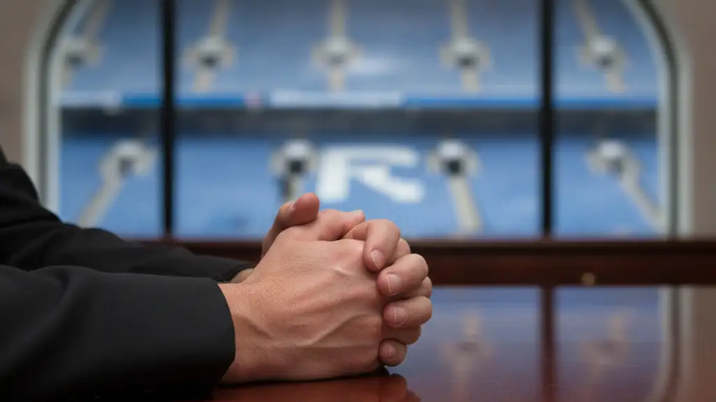 Manager's hands clasped on a desk, blurred stadium in background.