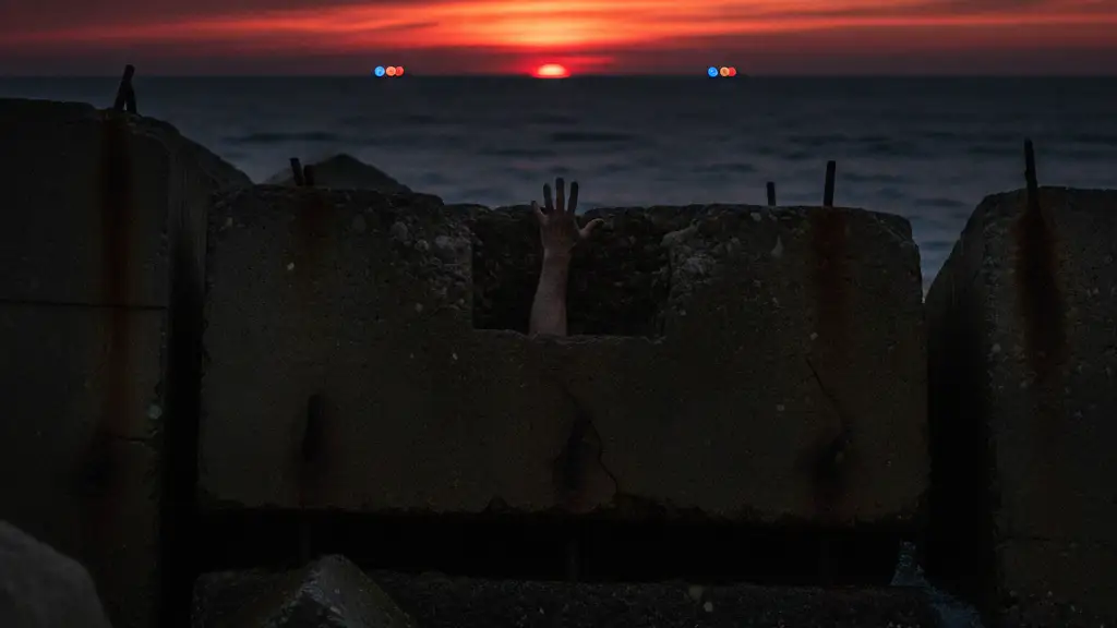 A lone hand reaching over a dark sea barrier at dusk, silhouetted against the setting sun.