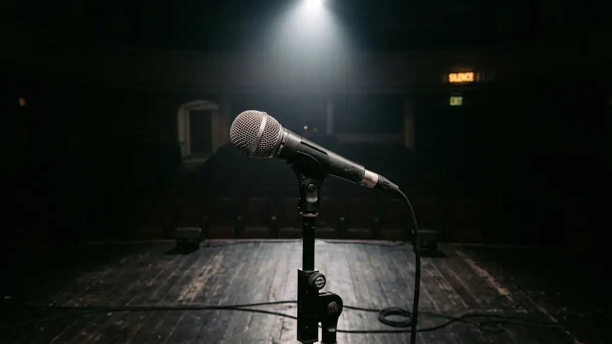 A single studio microphone stands under a spotlight on an empty, dark stage.