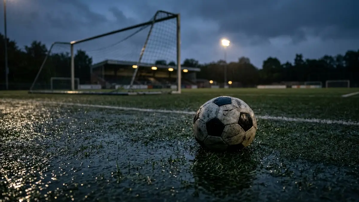 Deflated soccer ball on a wet, dimly lit football pitch at dusk, goal net blurred.