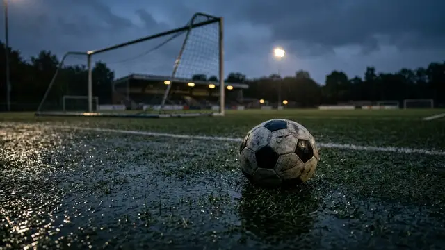 Deflated soccer ball on a wet, dimly lit football pitch at dusk, goal net blurred.