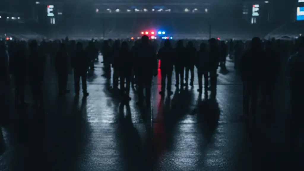 Blurred crowd outside stadium with police tape and emergency lights reflecting on wet ground.