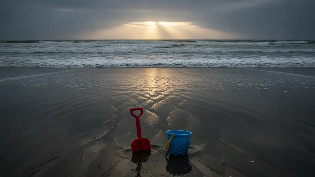 A child's sand shovel and bucket half-buried in wet sand by a turbulent ocean.