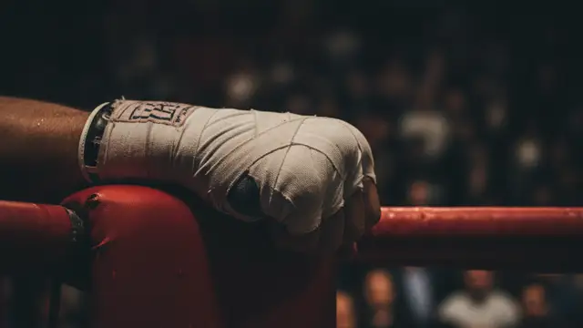 A gloved fist rests on a red corner post in a dimly lit boxing ring.