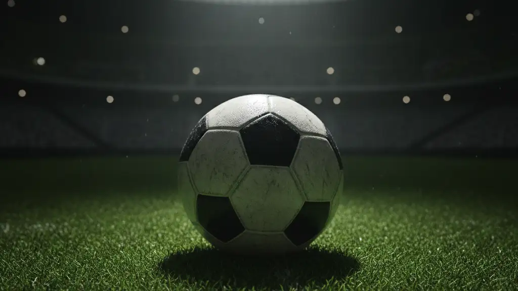 A football on a green pitch under a spotlight, stadium blurred in background.