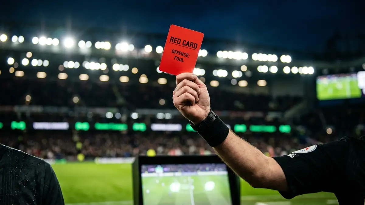 A referee's hand holds up a red card in a stadium under bright floodlights.