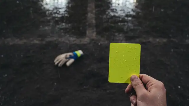 Referee's hand holding a yellow card, a goalkeeper's glove on a wet pitch.
