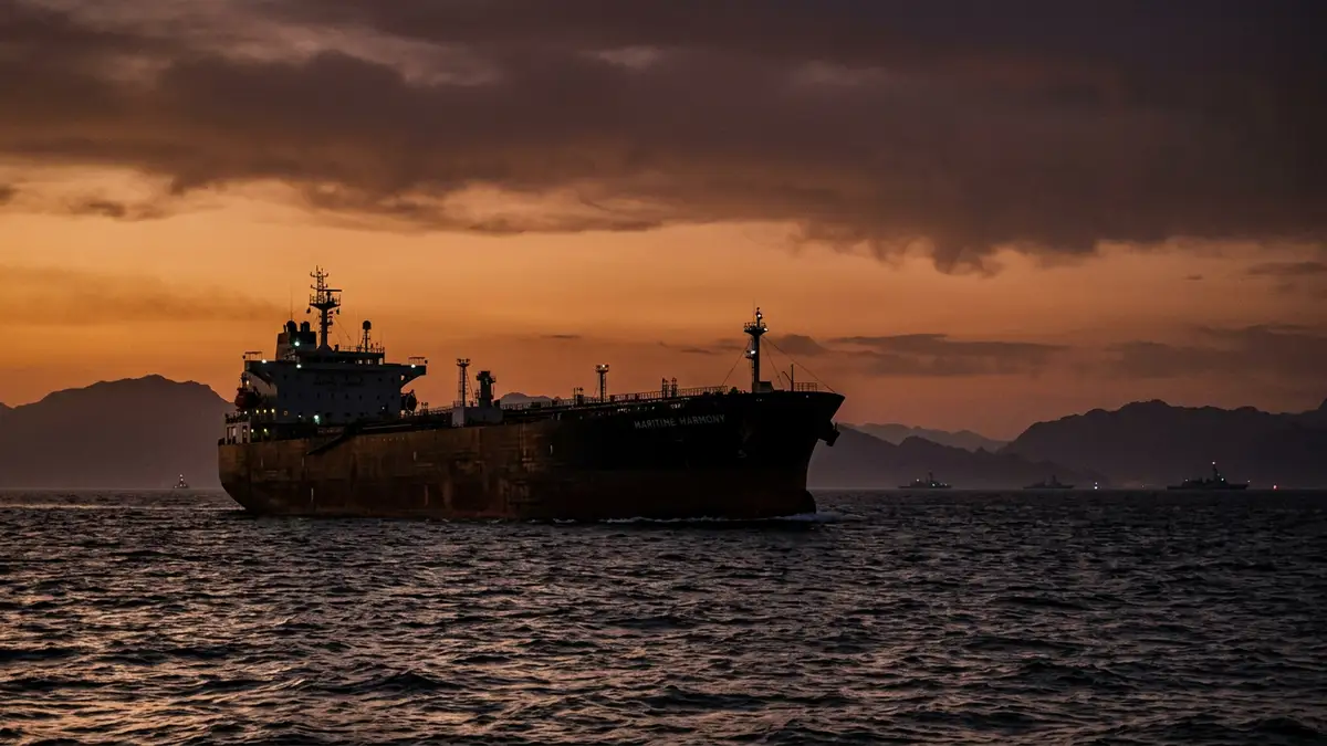 A large oil tanker silhouette at sunset in the Strait of Hormuz with naval ships distant.