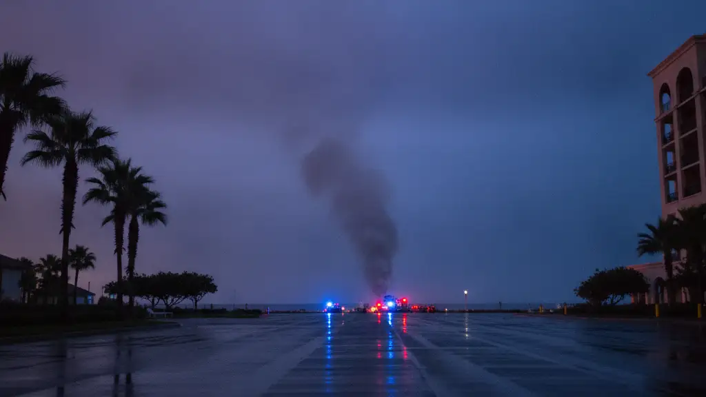Emergency lights reflect on wet pavement with distant smoke and palm tree silhouettes.