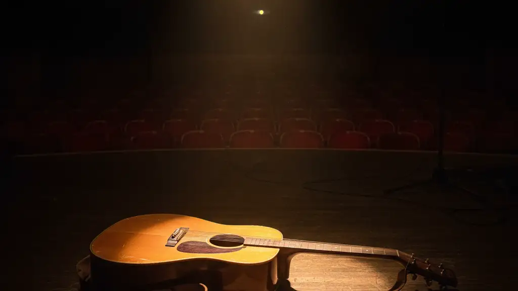 A lone vintage acoustic guitar on an empty, dimly lit stage under a single spotlight.