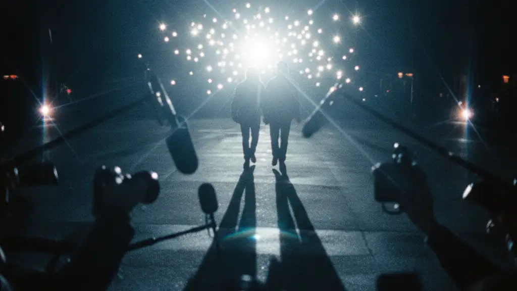 Blurred silhouettes of two men walking towards a blinding flash of paparazzi cameras.