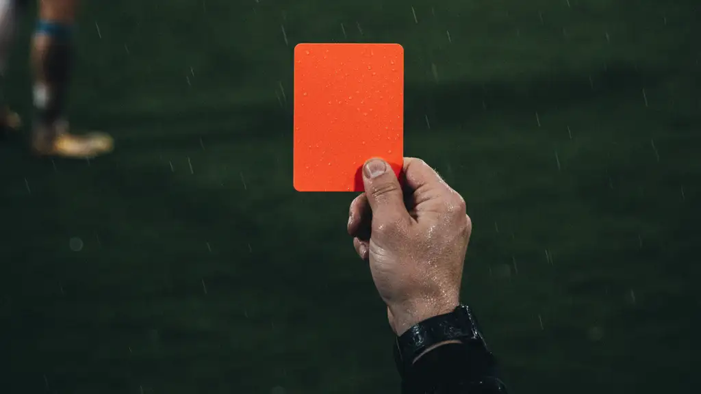 Referee's hand holding a red card on a football pitch, blurred background.