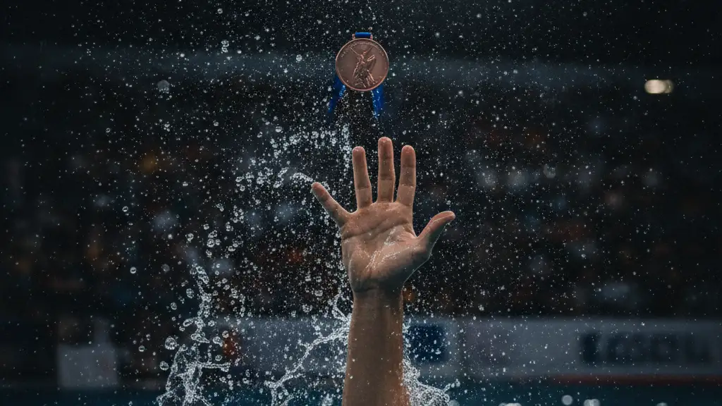 Swimmer's arm reaching for a bronze medal, face not visible, blurred stadium background.