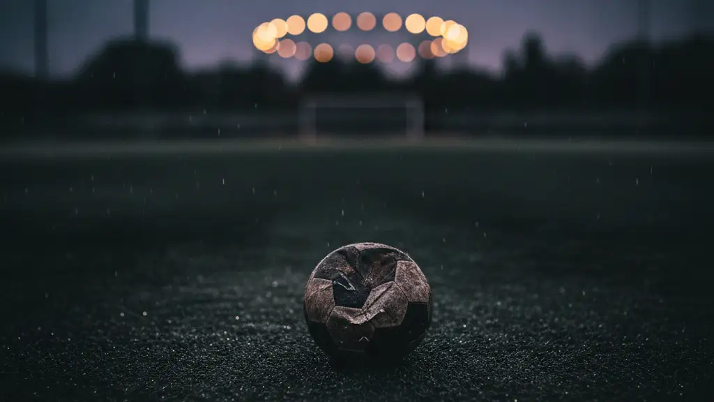 Deflated football on a wet, empty pitch under stadium lights at dusk.