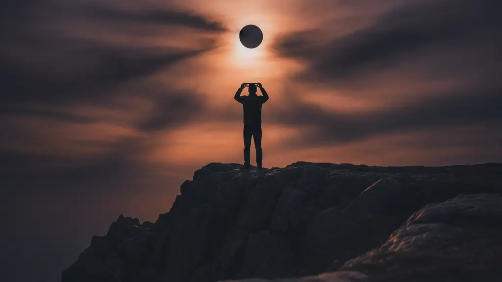 Silhouetted figure on cliff edge holding eclipse glasses, looking at a partially obscured sun.