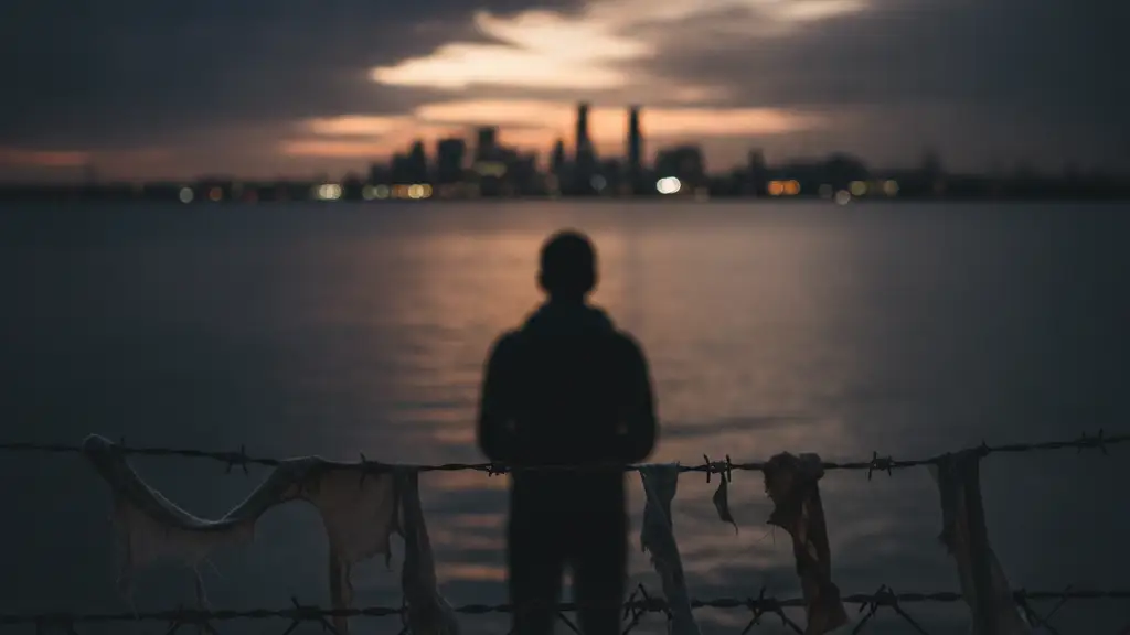 Silhouetted figure at a barbed wire fence looking towards a distant city.