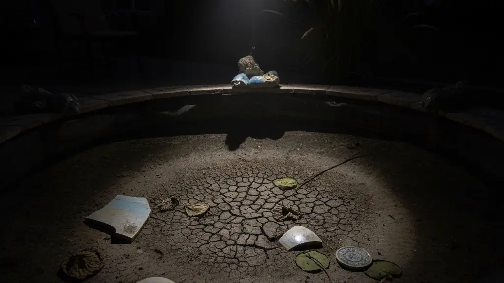Shattered, empty koi pond with broken ceramic and dry lily pads under dramatic spotlight.