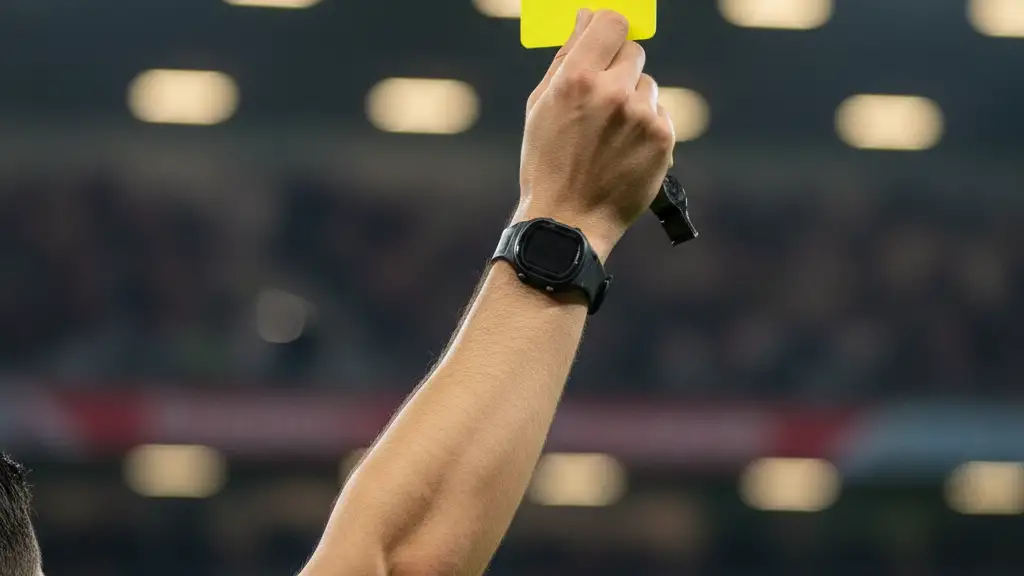 A referee's hand holds up a yellow card in a blurred, brightly lit stadium.