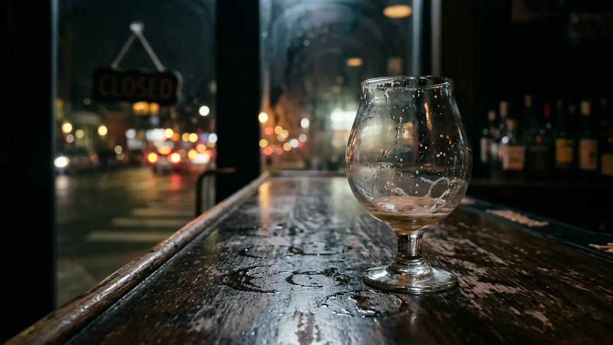 An empty beer glass sits on a dark wooden bar in front of a blurred window.