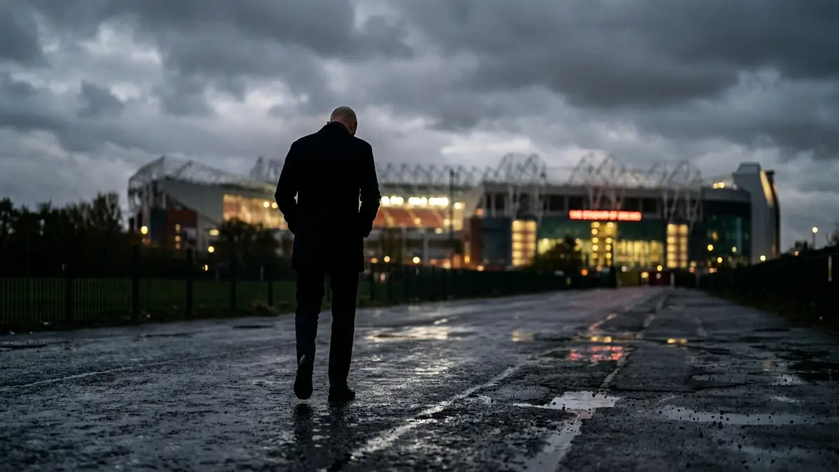 Silhouetted football manager walks away from a stadium under an overcast sky.