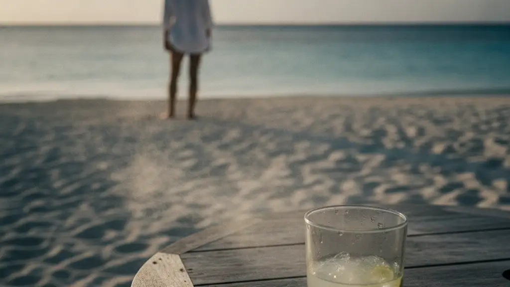 Half-empty cocktail glass on a beach table, blurred figure in straw hat looking at ocean.