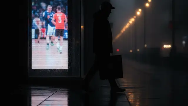 Silhouette of a shopper with a bag, passing a store reflecting a sports event.