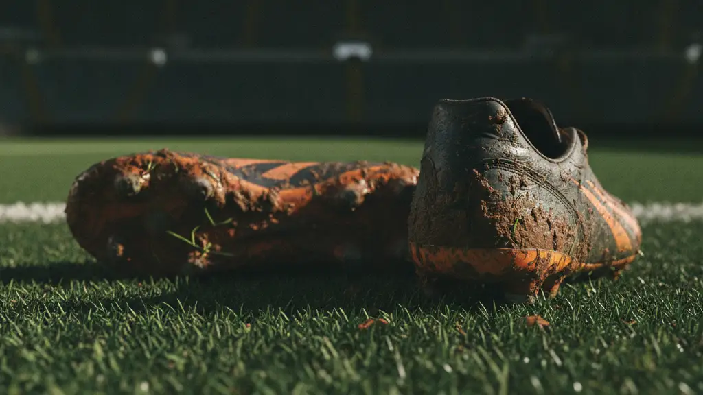Mud-splattered soccer cleats on a green pitch, blurred stadium scoreboard in background.