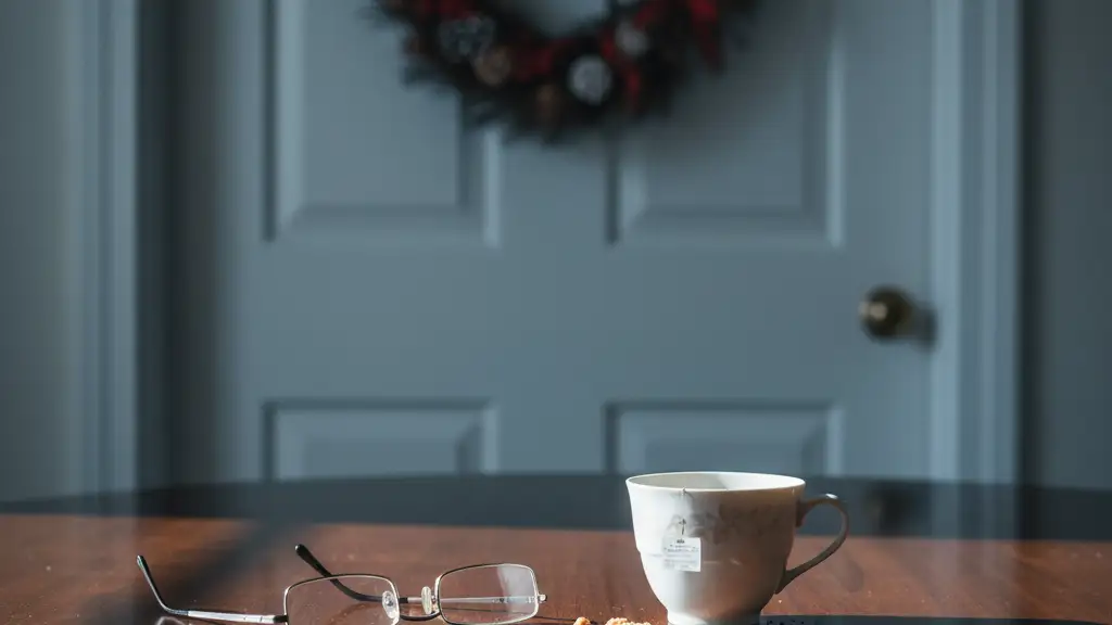 A teacup and reading glasses on a wooden table with a blurred Christmas wreath background.