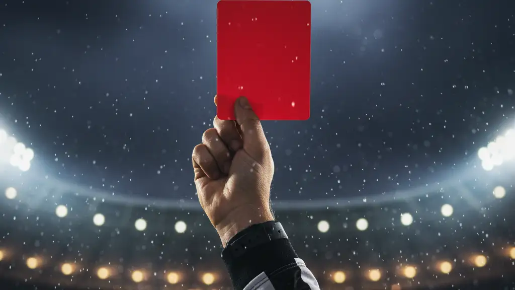 A referee's hand holding up a red card in a rain-slicked football stadium at night.