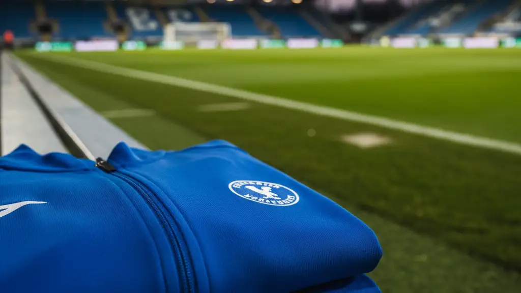 A blue team jacket sits on an empty bench beside a professional football pitch.